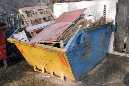 The image shows a large, industrial-sized metal skip in an outdoor area, painted in yellow on the front and blue on the sides, with visible signs of rust and weathering. The skip is filled with assorted waste materials, including wooden pallets, pieces of wood, metal sheets, and various discarded packaging, some leaning against the sides and others piled haphazardly inside. The waste items vary in texture from smooth painted surfaces to rough, splintered wood and corrugated metal. The skip is positioned on a paved, slightly damp surface near a white wall and a wooden fence, suggesting a commercial or industrial setting. The surroundings include a small section of fencing and other items indicative of a site preparing for rubbish removal services, possibly handled by a local waste management company like Office Clearance Heston, servicing the Heston area with commercial clearance and rubbish removal. Natural daylight illuminates the scene, casting minimal shadows, highlighting the weathered appearance of the waste and the skip's metal surface.