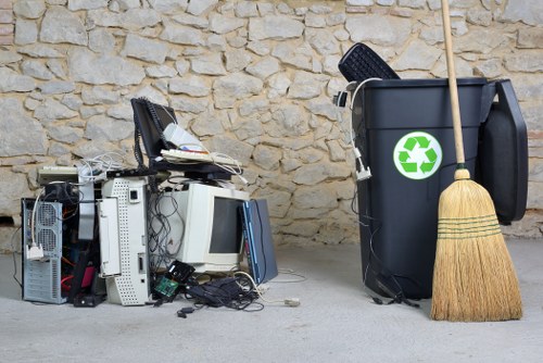 A pile of discarded electronic waste, including old computer towers, monitors, keyboards, and various cables, is positioned on a concrete surface against a textured beige stone wall. To the right, there is a large black rubbish bin with a green recycling symbol on its side, indicating it is designated for waste collection, alongside a wooden-handled broom leaning against it. The scene appears to be outdoors, possibly outside a commercial property in Heston, with natural lighting highlighting the materials' textures and colors, which range from the metallic and plastic finishes of the electronic devices to the rough stone wall background. The arrangement suggests an ongoing or recent electronic waste disposal effort, with a focus on responsible rubbish removal services offered by Office Clearance Heston.