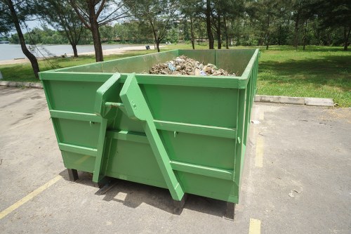 A large green metal skip container filled with mixed waste materials including cardboard, plastic, and general rubbish, situated on a paved area within an outdoor park-like setting. The skip is positioned on a parking lot, with the surrounding environment featuring a grassy area, trees, and a body of water visible in the background. The skip has a sturdy construction with horizontal reinforcement ridges, and the waste inside appears loosely piled, extending to the top edges. The scene is illuminated by natural daylight, highlighting the vibrant green of the skip and the earthy tones of the waste, indicative of a professional rubbish removal service such as Office Clearance Heston operating in the Heston area, near London.