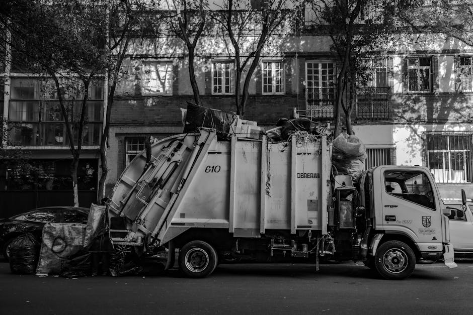 A white waste collection truck with the identification number 610 and the label 'Dorebai' on its side is parked on a city street in front of a multistory brick building with white-framed windows. The